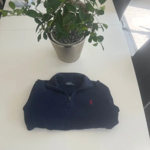 Marinblå half zip tröja Polo Ralph Lauren - Marinblå långärmad tröja från Polo Ralph Lauren med half zip och röd broderad logga på bröstet. Tröjan har ribbade muddar och krage med vit och röd rand på baksidan. Tillverkad i mjuk bomull, perfekt för dig med en bra stil. Säljes billigt med tanke på fläkarna längst ner på tröjan.