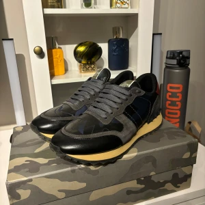 Valentino Garavani Rockrunner - Snygga Valentino Garavani Rockrunner sneakers i svart, grått och mörkblått med coolt kamouflagemönster. Skorna har detaljer i mocka och skinn, samt en beige sula och klassisk snörning. Perfekta för dig som vill sticka ut med exklusiva och trendiga sneakers.