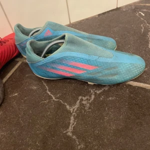 Blå fotbollsskor från Adidas - Säljer ett par blå fotbollsskor från Adidas med rosa detaljer och klassiska tre ränder på sidan. Skorna har en strömlinjeformad design utan snörning och ovandel i syntetmaterial. Yttersulan har fasta dobbar för bra grepp på planen.
