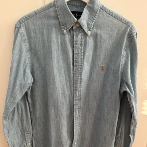 Ljusblå skjorta från Ralph Lauren - Klassisk ljusblå skjorta i slim-fit modell från Ralph Lauren i bomull med broderad logga på bröstet. Skjortan har långa ärmar och knäppning framtill. 