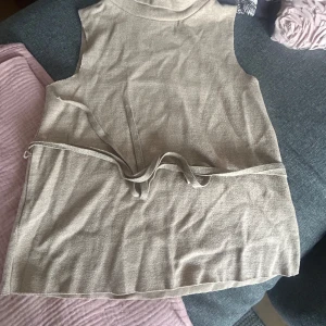 Beige stickad väst från Zara med knyt - Säljer en beige stickad halter topp från Zara i storlek M. Tröjan är ärmlös, har rund halsringning och ett matchande knytband i midjan för en snygg siluett. Bra skick!