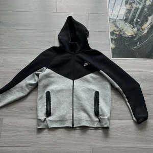 Nike tech kofta - Nike kofta i svart och ljusgrått med snygg kontrastdesign. Koftan har huva, hel dragkedja framtill och två fickor med svarta dragkedjor. Perfekt för chill dagar eller när du vill ha en sportig look. Nike-logga på bröstet och mjukt material.