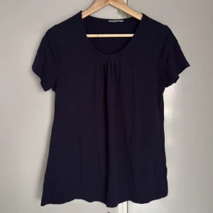 Mörkblå kortärmad blus i bomull - Enkel och stilren mörkblå blus med korta ärmar och rund halsringning. Framtill finns små veck som ger en snygg detalj. Tillverkad i mjuk bomull, perfekt för en clean och avslappnad look. Tvättlapp och storleksklass bortklippta