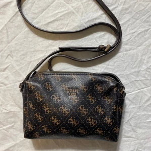 Svart axelväska från Guess med monogram - Snygg svart axelväska från Guess med brunt monogramtryck över hela väskan och guldfärgad logga framtill. Väskan har justerbar axelrem och dragkedja upptill. Tillverkad i syntetmaterial med lyxig känsla och stilren form.