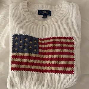 Vit stickad tröja Polo Ralph Lauren - Vit stickad tröja från Polo Ralph Lauren med en stor amerikansk flagga broderad på bröstet. Tröjan har rund halsringning och är gjord i grovstickad bomull, perfekt för dig som gillar klassisk och preppy stil.
