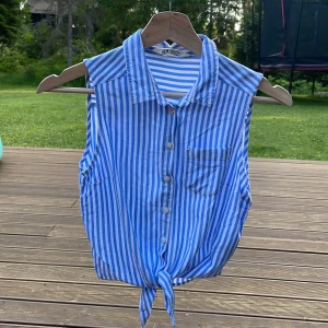 Blåvit randig ärmlös blus H&M - Säljer en blå och vit randig ärmlös blus från H&M. Toppen har klassisk skjortkrage, knappar framtill, bröstficka och knytning nedtill. Perfekt för varma dagar och ger en chill sommarvibe.                                  Den är lite liten i storleken skulle själv säga att den var ca 140-148.