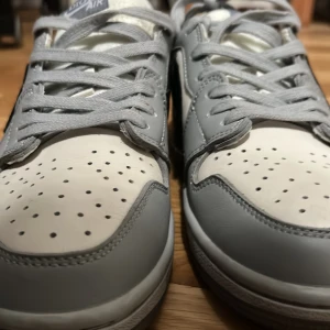 Nike Air Jordan 1 Low grå/vit - Snygga Nike Air Jordan 1 Low sneakers i grått och vitt skinn med klassisk svart swoosh på sidan. Skorna har perforerad tå, platt sula och grå snörning. Perfekta för dig som vill ha en stilren och ikonisk sneaker med streetkänsla.