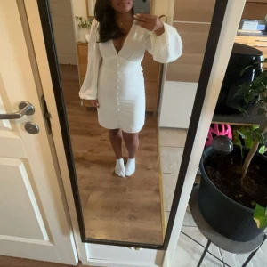 Vit kortklänning från NA-KD - Superfin vit kortklänning från NA-KD med v-ringning, långa puffärmar och knappar framtill. Klänningen har diskret prickigt mönster i tyget och smock i ryggen för en snygg passform. Perfekt för dig som vill ha en romantisk och trendig look. Den är i en väldigt bra skick då jag bara har använt den vid två tillfällen.💗