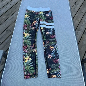 Blommiga leggings från Stronger XS - Färgglada leggings från Stronger i storlek XS med tropiskt blommönster och vita detaljer vid låret. Hög midja och stretchigt material i polyester och elastan. Perfekta för träning eller när du vill sticka ut lite extra.