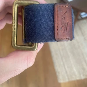 Mörkblå Polo Ralph Lauren bälte - Snyggt mörkblått bälte från Polo Ralph Lauren med robust mässingsfärgat spänne och detaljer i brunt skinn. Bältet är gjort i bomullstyg och har en klassisk, stilren look med Polo-logga på läderpatchen. Perfekt accessoar för att lyfta din outfit. Haft det i några år och tyvärr blivit för tjock för det nu. 