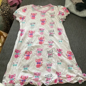 Söt kattklänning från Lindex - Supersöt kort klänning från Lindex med färgglada kattmotiv i rosa, turkos och grått. Klänningen har volangkant nertill, korta puffärmar och en liten rosett vid halsen. Perfekt för dig som älskar gulliga prints och mjuka material.