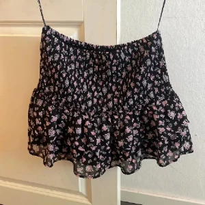 Blommig kortkjol från Jennyfer - Supersöt svart kortkjol från Jennyfer med små rosa och vita blommor. Kjolen har smockad midja och volang nertill som ger en flowy känsla. Perfekt för dig som gillar romantiska vibes och vill ha något lätt och luftigt till sommaren.