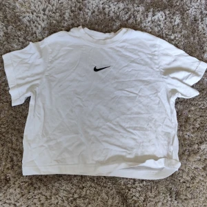 Vit Nike t-shirt med svart logo - Vit t-shirt från Nike med klassisk svart swoosh-logga på bröstet. Enkel och clean design med rund hals och korta ärmar. Tillverkad i mjuk bomull som känns skön mot huden. Perfekt för en sportig och avslappnad stil.
