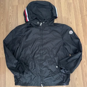 Svart vindjacka från Moncler - Svart vindjacka från Moncler med huva och dragkedja framtill. Jackan har Moncler-logga på ärmen och snygga röd-vita ränder på huvan. Tillverkad i lätt polyester, perfekt för blåsiga dagar. Klassisk passform och elastiska muddar vid ärmslut.