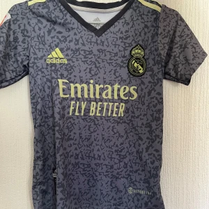 Real Madrid Benzema Adidas matchställ - Säljer ett Real Madrid matchställ från Adidas med Benzema och nummer 9 på ryggen. Tröjan är kortärmad, grå med svart mönster och gula detaljer, och har klubbmärke och sponsortryck. Shortsen är svarta med gula ränder och klubbmärke. Materialet är lätt och andas.