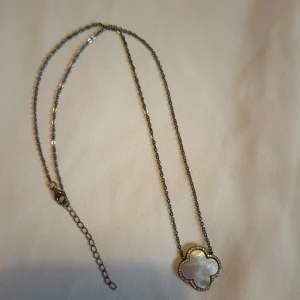 Halsband med fyrklöver i pärlemor SILVER  - Snyggt halsband med tunn silverfärgad kedja och ett fyrklöverformat hänge i vit pärlemor, omgiven av små glittrande stenar. Perfekt för dig som gillar stilrena och eleganta smycken med en touch av lyx.