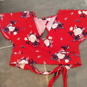 Röd blommig omlottopp med knyt - Superfin röd omlottopp från Boohoo med vita och blå blommor. Toppen har korta ärmar, v-ringning och knyt i midjan. Perfekt för sommarens alla tillfällen och ger en riktigt fräsch vibe med sitt blommiga mönster och öppna rygg.