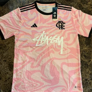 Rosa Flamengo x Stüssy fotbollströja Adidas - Unik rosa fotbollströja från Adidas i samarbete med Flamengo och Stüssy. Tröjan har korta ärmar, svart krage och svarta detaljer på axlarna. Mönstret är swirligt i rosa och vitt, med stora Stüssy-loggan framtill. Tillverkad i lätt och ventilerande material.