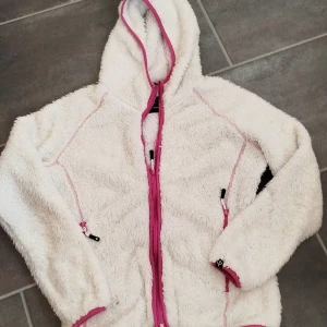 Vit teddyjacka från Cross Sportswear - Mysig vit teddyjacka med huva från Cross Sportswear. Jackan har rosa dragkedja och detaljer, samt två fickor med dragkedja. Perfekt för kyliga dagar när du vill vara både varm och snygg. Långärmad och fluffig med sportig känsla.