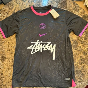 PSG x Stüssy x Nike svart fotbollströja - Svart PSG fotbollströja i samarbete med Stüssy och Nike. Tröjan har korta ärmar, rosa detaljer vid krage och ärmslut, samt ett subtilt blommönster över hela tyget. Stor Stüssy-logga framtill och Paris Saint-Germain-emblem i rosa. Tillverkad i Dri-FIT-material.