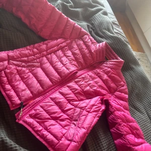 Rosa och orange dunjacka från Tuxer - Säljer en färgstark dunjacka från Tuxer i rosa med orange insida. Jackan är quiltad, har dragkedja framtill och två fickor. Perfekt för dig som vill sticka ut med en cool och sportig look. Passformen är normal och jackan är lätt och smidig att bära.