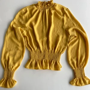 Gul blus med volang från NLY Trend - Superfin gul blus från NLY Trend med smockad volangkrage, långa puffärmar och smockade muddar. Blusen har dragkedja i nacken och markerad midja med volangkant. Perfekt för dig som vill sticka ut med färg och snygga detaljer.