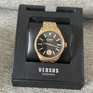 Guldig Versus Versace klocka - Snygg klocka från Versus Versace med guldig länk och svart urtavla. Klockan har klassisk rund form, detaljer i guld och Versus-logga med lejonmotiv på urtavlan. Kommer i originalbox med garantikort och förpackning. Perfekt accessoar för att lyfta din stil.