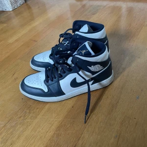 Nike Air Jordan 1 Mid Navy/Vit - Snygga Nike Air Jordan 1 Mid sneakers i marinblått och vitt skinn. Klassisk high-top siluett med perforerad tå, ikoniska Jordan Wings-loggan på sidan och svart swoosh. Skorna har snörning och en platt sula för en clean och sportig vibe.