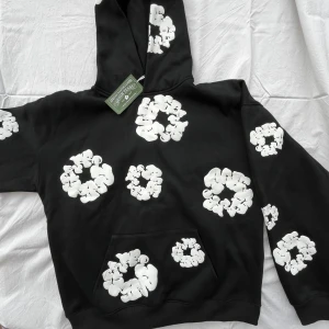 Svart hoodie med vita blommor Denim Tears - Säljer en svart hoodie från Denim Tears med stora vita blommönster över hela tröjan. Tröjan har huva, magficka och är tillverkad i 100% bomull. Perfekt statement-plagg för dig som gillar streetwear och unika prints.