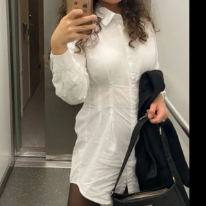 Skjortklänning  - Klassisk vit skjortklänning i bomull med lång ärm och knappar hela vägen framtill. Skjortan har en figurnära passform och krage, perfekt att styla oversized eller som klänning. Enkel och stilren design som funkar till många olika looks.