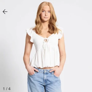 Vit blus med volangärm och knyt - Supersöt vit blus med volangärmar och knyt fram i urringningen. Toppen har en lätt rynkad, luftig passform och är något croppad. Perfekt till jeans eller kjol för en fräsch sommarlook.