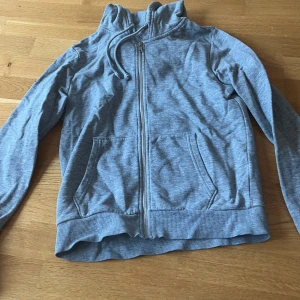 Grå hoodie med dragkedja från H&M - Mysig grå hoodie från H&M med hel dragkedja, huva med snörning och två stora fickor framtill. Tillverkad i mjuk bomullsblandning, perfekt för chill dagar. Klassisk passform och ribbade muddar vid ärmslut och nederkant.
