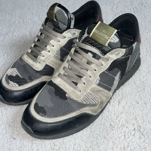 Valentino Rockrunner  - Snygga Valentino Rockrunner sneakers med grå och svart camouflage-mönster, detaljer i mocka och mesh samt svarta och vinröda skinnpartier vid hälen. Klassisk snörning och platt sula. Perfekta för dig som vill sticka ut med exklusiva och trendiga skor. Lite smutsiga men annars är dom i fint skick. Hör av dig vid minsta fundering!💫