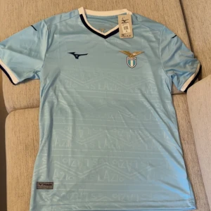 Ljusblå Lazio fotbollströja - Snygg ljusblå fotbollströja med SS Lazio-logga på bröstet. Tröjan har korta ärmar, V-ringning med vit och mörkblå kant samt diskret mönster med Lazio-text i tyget. Perfekt för dig som älskar italiensk fotboll.