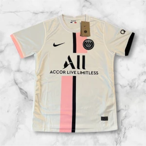PSG x Nike vit och rosa fotbollströja - Snygg PSG fotbollströja i vitt med en rosa och svart vertikal rand på framsidan. Tröjan har korta ärmar, svart Nike-logga och Paris Saint-Germain-emblem på bröstet. Materialet är lätt och andas, perfekt för match eller träning. Svart och rosa detalj på ärmsluten.