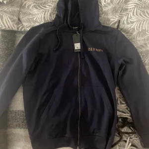 Mörkblå hoodie från Lyle & Scott - Säljer en snygg mörkblå hoodie från Lyle & Scott med dragkedja och huva. Tröjan har en liten gul logga på ärmen och text på bröstet. Perfekt för en avslappnad stil. Den är helt ny. Är 190 lång och tycker den är lite för stor.