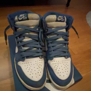 Nike Air Jordan 1 Retro High  - Säljer ett par Nike Air Jordan 1 Retro High OG i blått, grått och vitt. Skorna har klassisk snörning och en ikonisk design med Nike Air-logga på plösen. Skorna är laced och det tillkommer vita skosnören.
