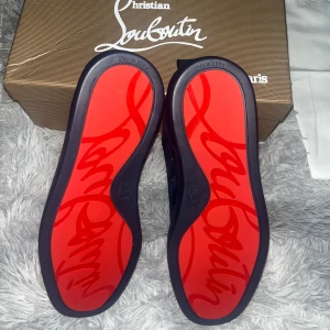 Mörkblå sneakers från Christian Louboutin - Snygga mörkblå sneakers i mocka från Christian Louboutin med den ikoniska röda sulan. Skorna har snörning och en rund tå, vilket ger en stilren look. Perfekta för den som vill ha en touch av lyx i vardagen.