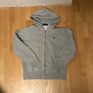 Grå hoodie från Ralph Lauren - Säljer en stilren grå hoodie från Ralph Lauren med dragkedja och huva. Storlek M men passar perfekt 170-175. Tröjan har långa ärmar och en liten broderad logga på bröstet. Liten grå fläck på baksidan av armen. 