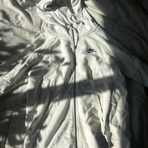 Vit hoodie från Nike - Säljer en stilren vit hoodie från Nike med dragkedja och huva. Perfekt för en avslappnad look. Den har långa ärmar och en liten Nike-logga på bröstet. Passar bra till både träning och vardag.