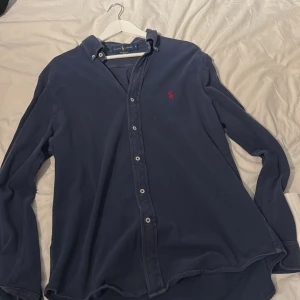 Blå skjorta från Ralph Lauren - Säljer en stilren blå skjorta från Ralph Lauren med klassisk krage och knappar framtill. Skjortan har en broderad röd logotyp på bröstet och är tillverkad i ett lätt och bekvämt material.