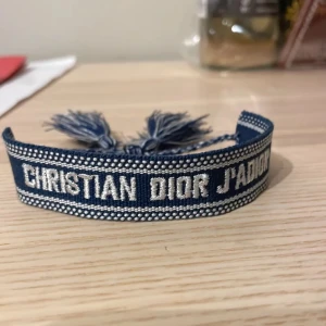 Blått armband från Christian Dior - Snyggt blått armband från Christian Dior med texten 'J'ADIOR' i vitt. Armbandet har en fransig detalj i ändarna som ger en bohemisk touch. Perfekt för att lyfta vilken outfit som helst!🔥🔥