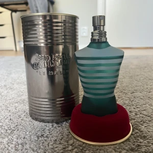 Jean Paul Gaultier Le Male Parfym - Ikonisk parfymflaska formad som en manlig torso med blå och grå ränder. Flaskan står på en röd bas och kommer i en metallburk med präglad text. En klassisk doft som är både djärv och tidlös.