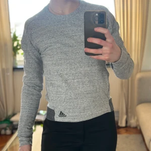 Grå långärmad tröja från Adidas - Säljer en grå långärmad sweatshirt från Adidas. Tröjan har en normal passform med muddar och en logga på framsidan. Perfekt till vardags eller träning!