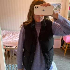 Svart dunväst från Canada Goose - Säljer en svart dunväst från Canada Goose. Västen har en klassisk design med knappar och dragkedja framtill. Perfekt för kyligare dagar när du vill hålla värmen utan att kompromissa med stilen. 