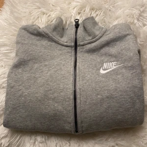 Grå hoodie från Nike - Säljer en stilren grå hoodie från Nike med dragkedja och logga på bröstet. Perfekt för en avslappnad look. Den har långa ärmar och en bekväm passform.