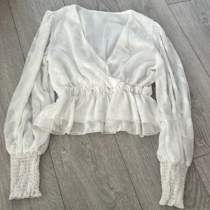 Vit blus med volangdetaljer - Säljer en elegant vit blus med v-ringning och volangdetaljer. Blusen har långa ärmar med smockade muddar och ett subtilt bladmönster. Perfekt för en stilren look.