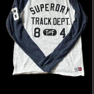 Vit och marinblå långärmad tröja från Superdry - Säljer en vit och marinblå långärmad tröja från Superdry med tryck 'Track Dept.' och siffrorna 8 och 4 på framsidan. Tröjan har en sportig stil med kontrasterande ärmar och rund halsringning.
