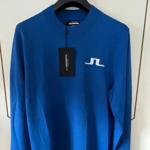 Blå golf tröja från J.Lindeberg - Snygg blå tröja från J.Lindeberg med ribbad krage och muddar. Den har en vit logotyp på bröstet och baksidan. Den är gjord av viskos och nylon vilket gör den stretchig och luftig.  Perfekt för en go vårdag eller sen sommarkväll ute på golfbanan men funkar även lika bra var som helst. Helt oanvänd med lappen kvar. Namn på plagget: ”Gus Knitted Sweater”. Slutsåld i princip överallt. Nypris ca 1500kr