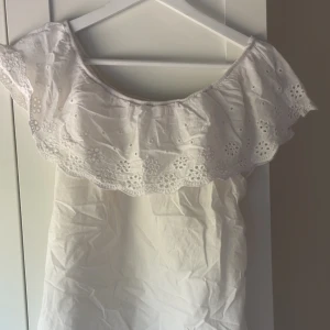 Vit offshoulder blus med spetsdetaljer - Säljer en söt vit offshoulder blus med spetsdetaljer. Perfekt för varma dagar med sin luftiga design och romantiska stil. Blusen har en vacker spetskant som ger en feminin touch.